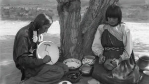 Two individuals in traditional attire sit by a tree, crafting pottery and baskets with various handcrafted items displayed between them.
