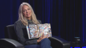 A person with long gray hair is seated in a dark room, holding up an open book featuring colorful collages.