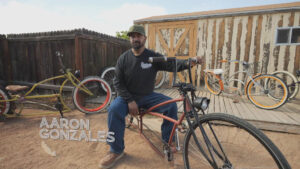 A man sits on a custom-built bicycle in a yard with two other bicycles and a wooden shed in the background. The name "Aaron Gonzales" is displayed on the image.