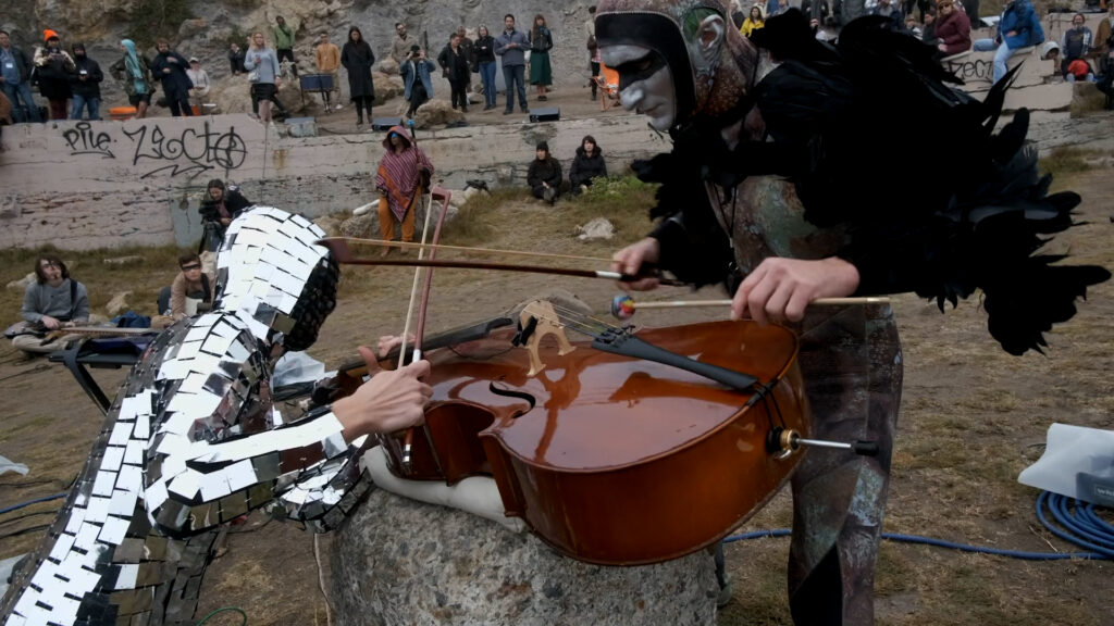 Two performers in costumes play a cello with a bow and a mallet in an outdoor setting, while a group of people watch in the background.