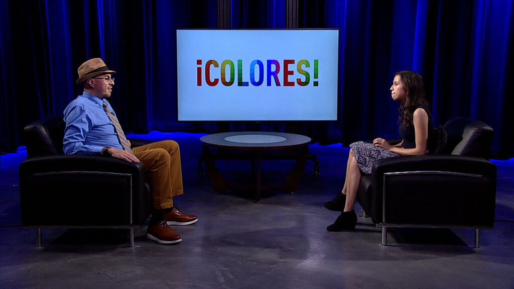 Two people are seated in armchairs on a dimly lit set, conversing. A large screen displaying "¡COLORES!" in colorful letters is positioned between them.