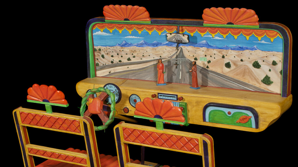 An artistic model of a bus dashboard with detailed painted scenery of a desert road and mountains. The model includes seats, a steering wheel, and small figurines in the background.