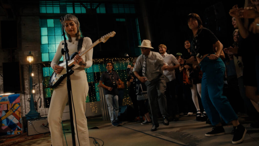 Musician plays electric guitar on stage while audience dances and claps in a dimly lit indoor venue.