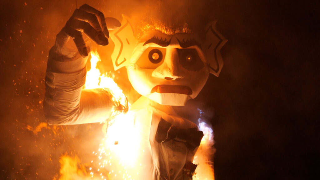 A large puppet with exaggerated features is engulfed in flames, creating a dramatic scene with sparks and smoke in the evening.