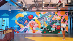 A woman stands in front of a vibrant mural depicting multiple faces, colorful snakes, and abstract patterns in a modern indoor setting.