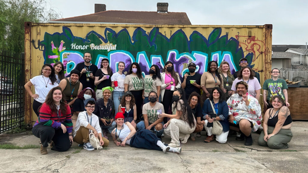 A diverse group of people poses in front of a colorful graffiti mural on a container, some holding drinks.