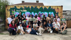 A diverse group of people poses in front of a colorful graffiti mural on a container, some holding drinks.