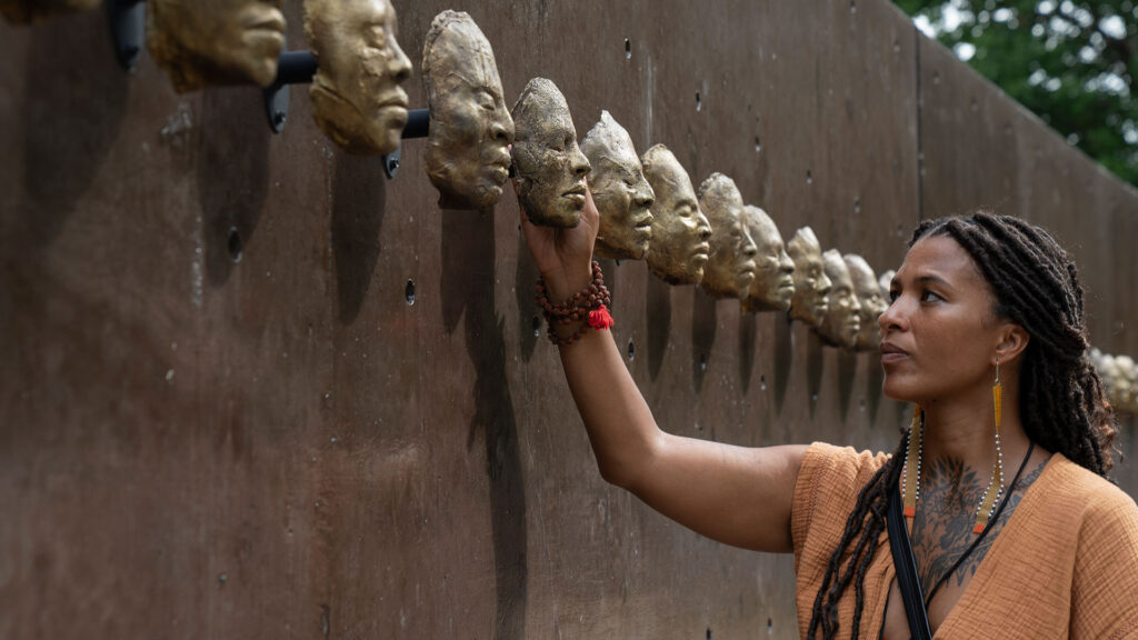 A woman touches a bronze face sculpture on a wall, part of a series of identical faces. She appears reflective, her hand carefully placed on one sculpture.