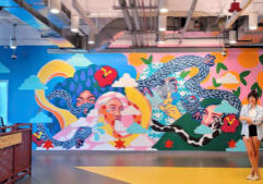 A woman stands in front of a vibrant mural depicting multiple faces, colorful snakes, and abstract patterns in a modern indoor setting.