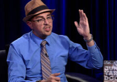 A man wearing a fedora, glasses, and a blue shirt with a striped tie is gesturing with his right hand while speaking. A "Junk Yard Dogs" logo is visible in the lower right corner.