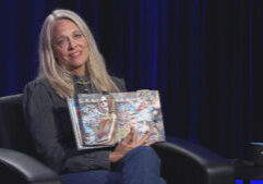 A person with long gray hair is seated in a dark room, holding up an open book featuring colorful collages.