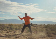 A man in an orange shirt flying a kite in the desert.