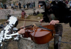 Two performers in costumes play a cello with a bow and a mallet in an outdoor setting, while a group of people watch in the background.