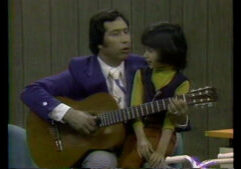 An adult in a purple jacket is sitting with a child, playing an acoustic guitar, while both look in the same direction.