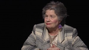 An older woman sitting at a table with a black background.