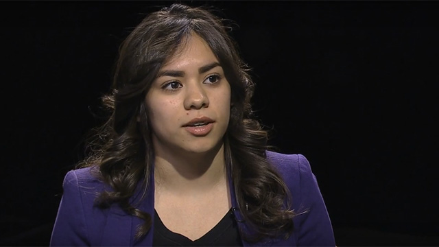 A woman in a purple jacket is talking to the camera.