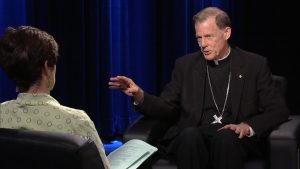 A priest is talking to a woman in a black chair.