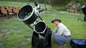 A scientist in outdoor clothing prepares a large telescope on grass.