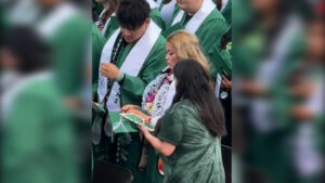A group of students in green graduation robes and white stoles are standing in a ceremony; two in the front are adjusting their stoles, and one is holding a decorated graduation cap.