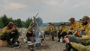 A group of firefighters in yellow uniforms sit around a campfire, eating and talking, with trees and a hazy sky in the background. Supplies and cooking pots are scattered around the campsite.