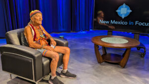 An elderly person with colorful attire sits in a chair on a studio set with a "New Mexico in Focus" sign on a screen next to them.