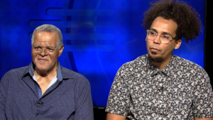 Two men are sitting side by side. The man on the left has gray hair and is wearing a blue shirt. The man on the right has curly hair, glasses, and is wearing a patterned shirt. The background is blue.