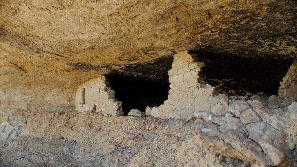 A partially collapsed ancient stone structure built into a rock overhang with visible remnants of walls, surrounded by rocky terrain and the distant sound of rushing water.