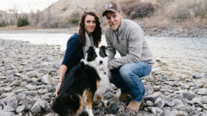 A woman and man kneel on a rocky riverbank with a black-and-white dog between them, the water glistening behind. They all look at the camera.