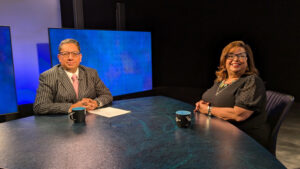 Two people sit at a studio table with blue backgrounds. Each has a dark mug in front of them. The man on the left is in a striped suit and tie, and the woman on the right is in a black dress.