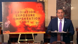 A man in a suit speaks at a podium beside a large sign that reads "Radiation Exposure Compensation Act" with images of people and an explosion in the background.