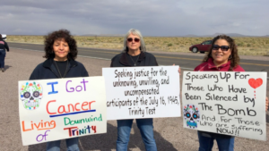 Three women are standing, holding signs that advocate for justice for those affected by the July 16, 1945 Trinity Test. The signs mention cancer and compensation for downwinders of the test.