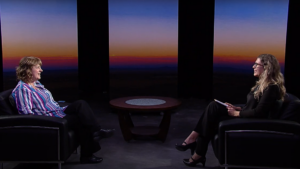 Two women seated and facing each other in armchairs during an interview, with the background featuring a gradient of sunset colors. A round table is placed in between them.