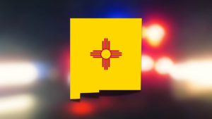 Image of the New Mexico state flag, a yellow field with a red Zia sun symbol in the center, set against a blurred background with red and blue lights.