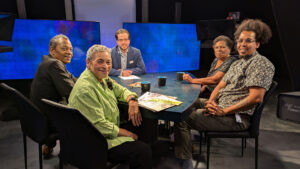 Five people sit around a table in a studio set up with blue background screens. The group includes two men and three women, one of whom has a newspaper on the table in front of her.