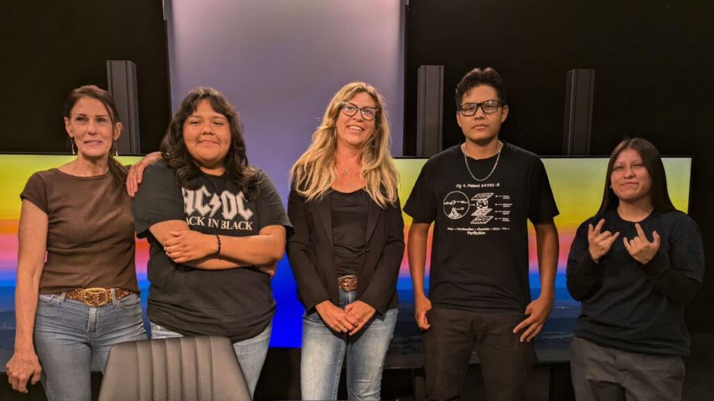 Five people standing side by side, smiling, with three women on the left and two men on the right, all wearing casual clothing. The background has computer monitors displaying gradient screens that flow like water.