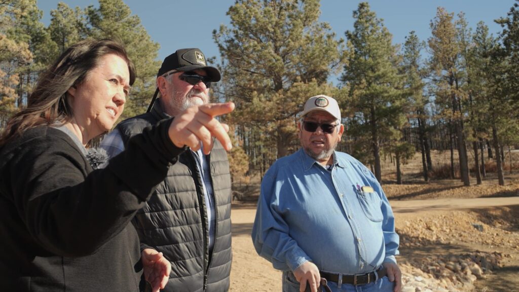 Three people stand outdoors near trees, engaged in conversation. The woman points to something off-camera while the two men wear hats and sunglasses. They are dressed in casual outdoor attire.