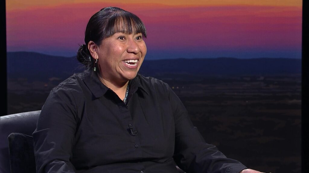 A person wearing a black shirt sits in front of a backdrop showing a colorful sunsetsky, smiling and speaking animatedly.