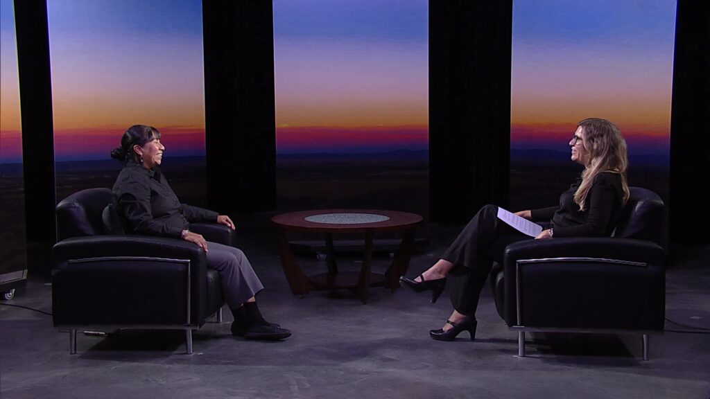 Two people sit in armchairs facing each other in a studio with a sunset backdrop. A round table is between them. One person is holding a sheet of paper.