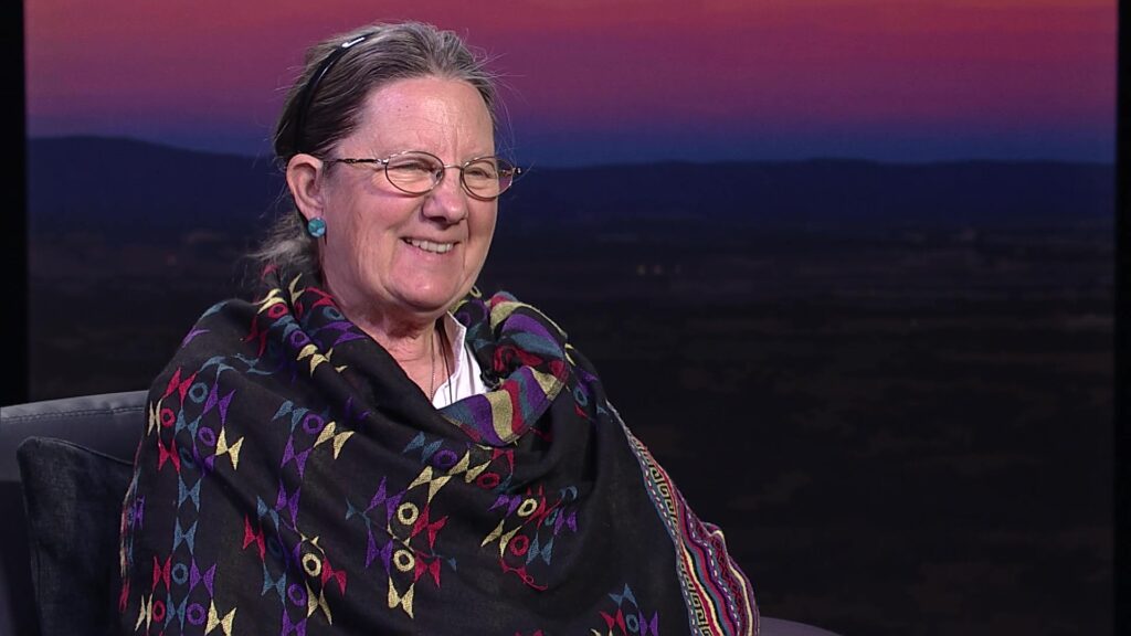 A smiling woman with glasses and gray hair, wearing a colorful patterned shawl, sits in front of a sunset backdrop with distant mountains.