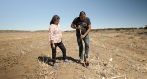 Two people working in a barren field under a clear sky. One person is digging with a shovel, while the other observes.
