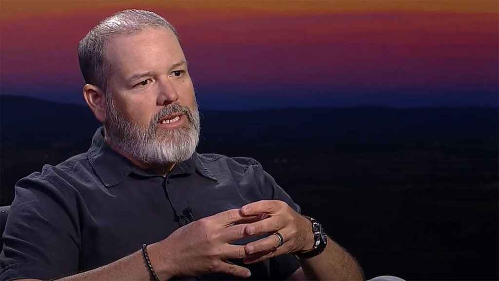 Man with gray beard and short hair talks while gesturing with his hands. He is wearing a dark shirt and sitting against a dusky outdoor backdrop.