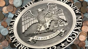 Close-up of the Great Seal of the State of New Mexico, featuring an eagle and the motto "Crescit Eundo," with assorted coins in the background.