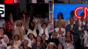 A group of people cheer at a public event, many smiling and raising their hands. A sign reading "New Mexico" is visible in the middle.