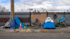 A row of tents, shopping carts, and personal belongings line a sidewalk in front of a barbed-wire fence, indicating a homeless encampment.