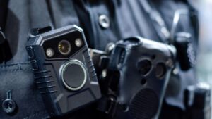 Close-up of a body-worn camera attached to a police officer's uniform, next to a radio and other law enforcement gear.