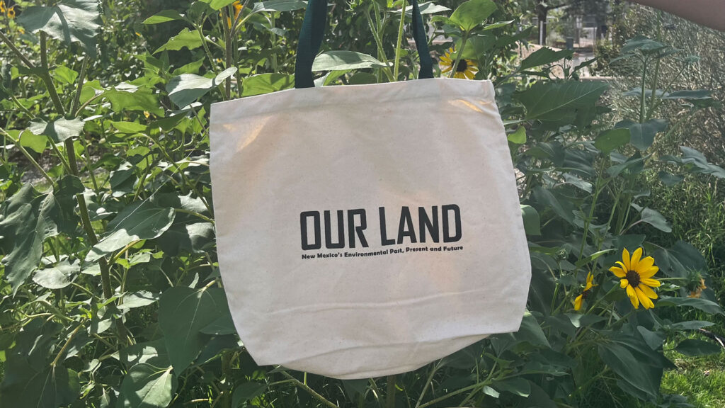 A beige tote bag with the words "OUR LAND: New Mexico's Environmental Past, Present and Future" printed on it, hanging among tall green plants and sunflowers, reflecting the vital importance of water in sustaining this vibrant landscape.