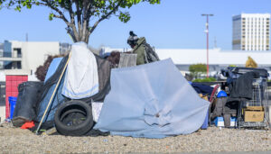 A makeshift shelter with a collapsed blue umbrella, various belongings, and a tire sits under a tree. Urban buildings and a parking lot are visible in the background.