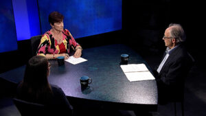 Three people are seated around a table in a studio, engaged in a discussion. Two women are facing each other, and one man is facing the camera. There are papers and coffee mugs on the table.