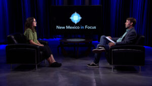 Two people sit across from each other in a studio with a "New Mexico in Focus" sign displayed on a large screen behind them, engaging in a conversation. The studio has blue lighting and dark curtains.