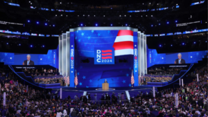 A large crowd attends the 2024 Democratic National Convention, with a speaker at the podium in front of a blue, patriotic-themed stage.